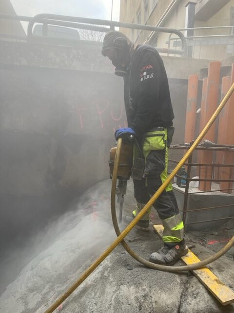 En byggarbetare med skyddsutrustning använder en tryckluftsdriven borrmaskin för spräckning av berg i ett byggschakt, omgivet av damm och betongväggar.